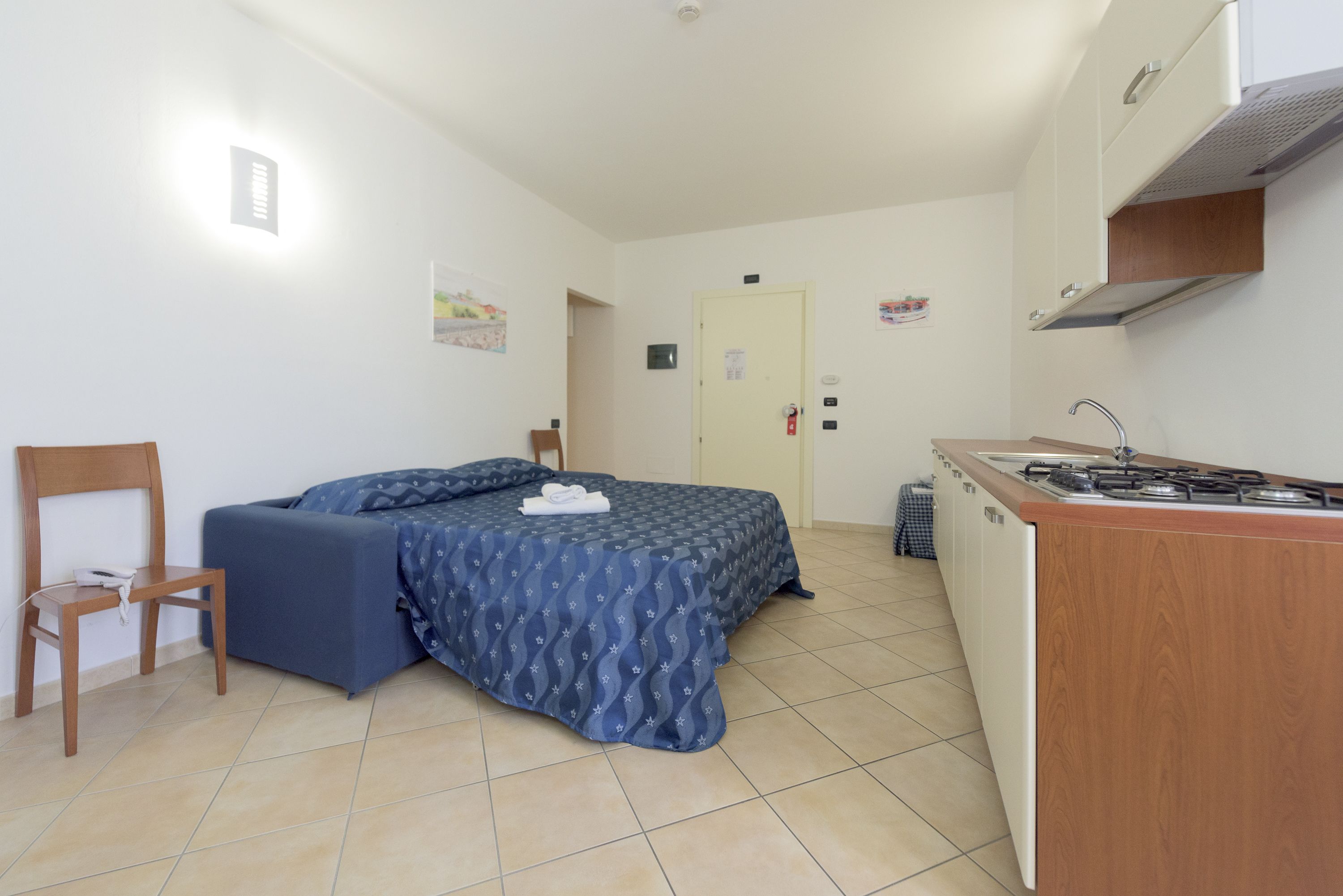 Quarto compacto com uma cama, uma pequena mesa, uma cadeira, um espaço de cozinha compacto e uma porta.