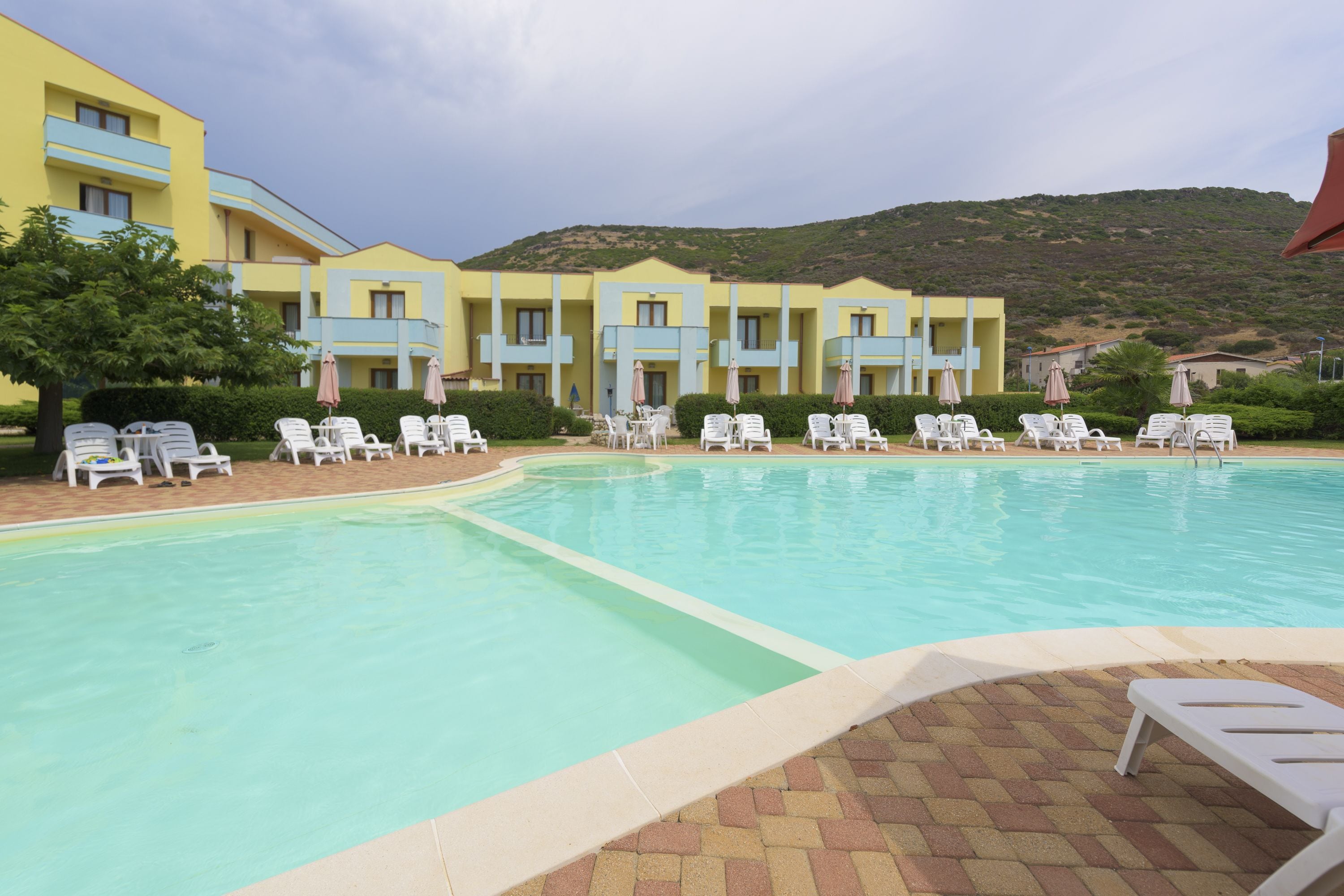 Una piscina con tumbonas y sombrillas, rodeada por un hotel con balcones azules y un paisaje montañoso al fondo.