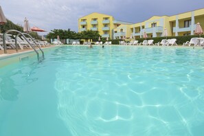 A swimming pool with clear water, surrounded by white lounge chairs and umbrellas, and a yellow building with balconies in the background. - Hotel Isola Rossa (Bosa)