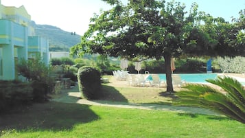 A poolside area with lounge chairs and umbrellas, surrounded by greenery and a building with a white facade.
