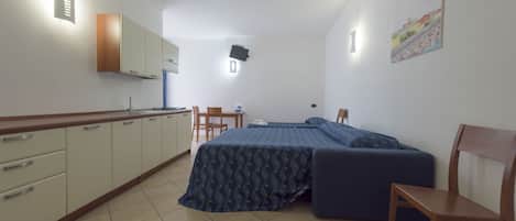 A compact room with a kitchenette, a bed with a patterned bedspread, and a chair.