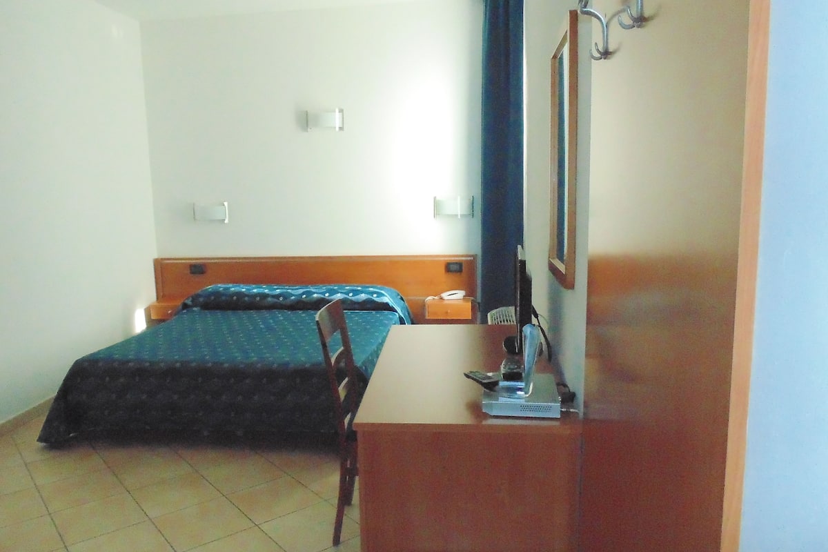 A hotel room with a bed, a chair, a desk with a computer, and a mirror.