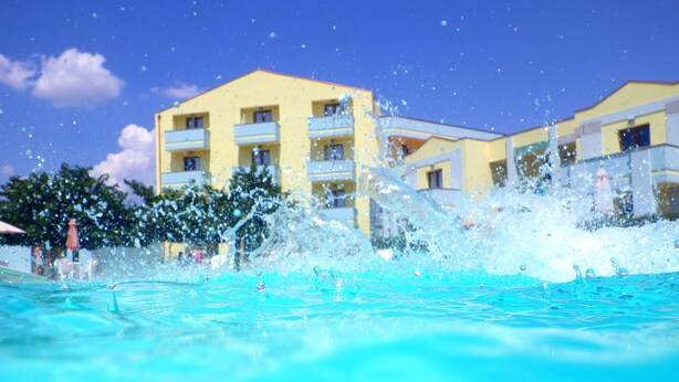 Una piscina con agua cristalina y salpicaduras, rodeada de edificios amarillos con balcones.