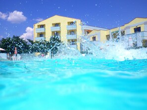 Piscina com água cristalina e respingos, rodeada por edifícios amarelos com varandas.