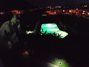 A nighttime view of a swimming pool with a green-lit water feature, surrounded by buildings and streetlights. - Hotel Isola Rossa (Bosa)