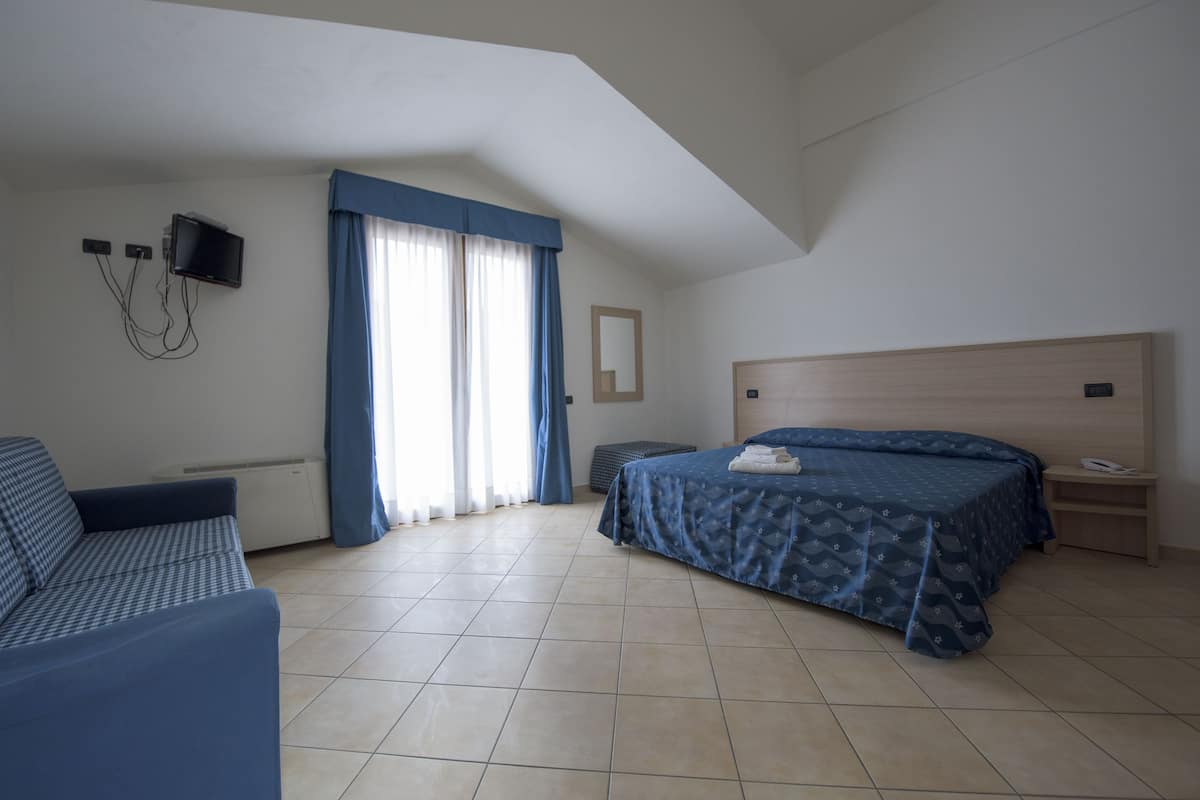 A bedroom with a bed, a sofa, a TV mounted on the wall, and a window with curtains.