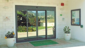 Entrada de hotel con puerta de vidrio, plantas en macetas y felpudo verde.