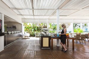 Un espacio de comedor al aire libre techado, con mesas de buffet, suelo de madera y vistas a la vegetación.