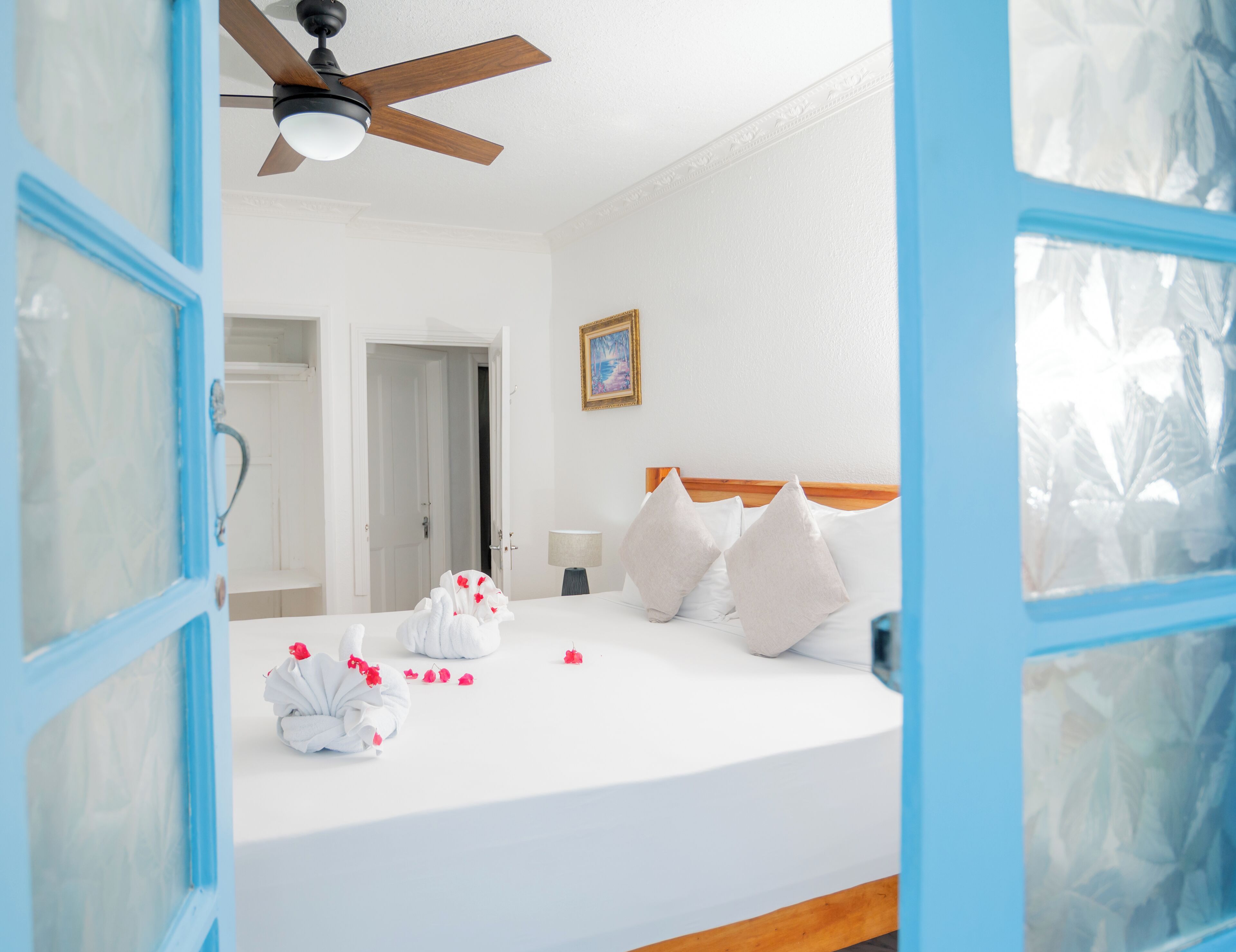 Une chambre d’hôtel avec un lit, un ventilateur de plafond, une porte et un tableau encadré au mur.