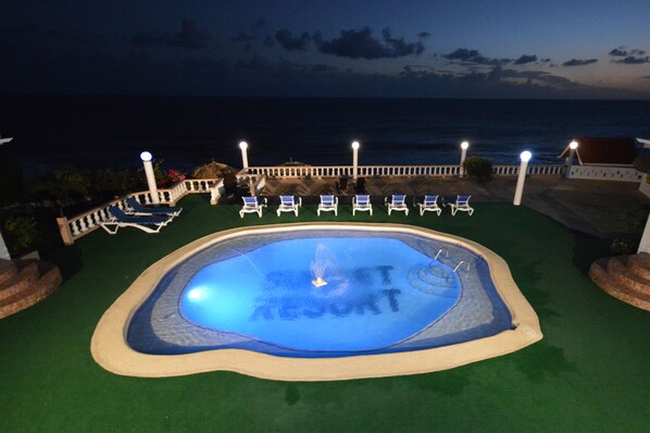 A swimming pool with a fountain at the center, surrounded by lounge chairs and a view of the ocean. - Beyond Sunset Resort & Villas (Treasure Beach)