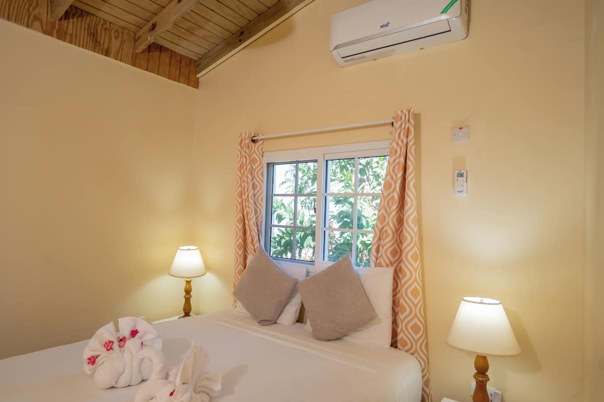 A hotel room with a bed, two bedside lamps, a window with curtains, an air conditioning unit, and a bedside table.