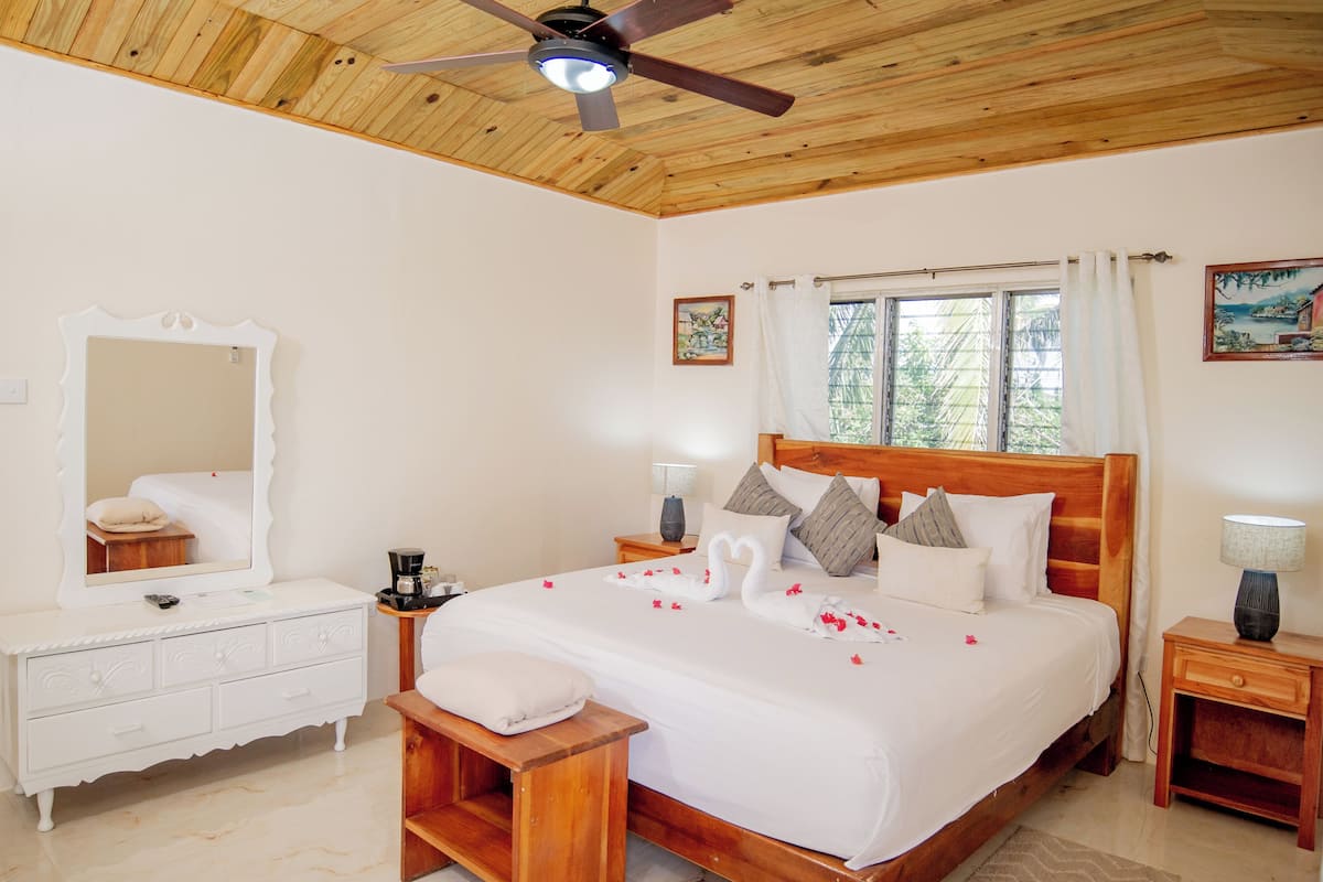 A bedroom with a wooden ceiling, a large bed with white bedding, a wooden nightstand, a bedside table with a lamp, and a mirror on the wall.