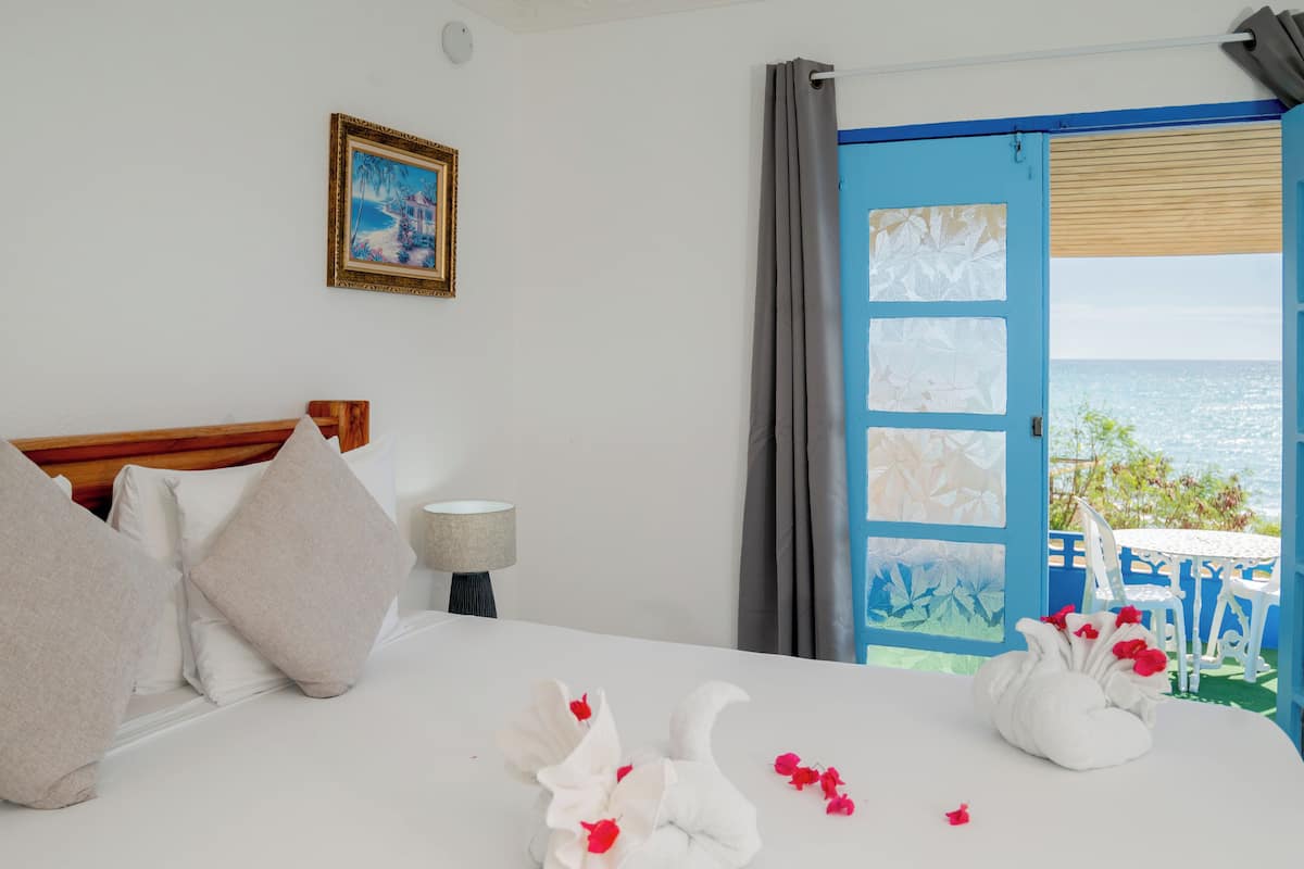A hotel room with a bed, a painting on the wall, a view of the sea through an open door, and a table with chairs outside.