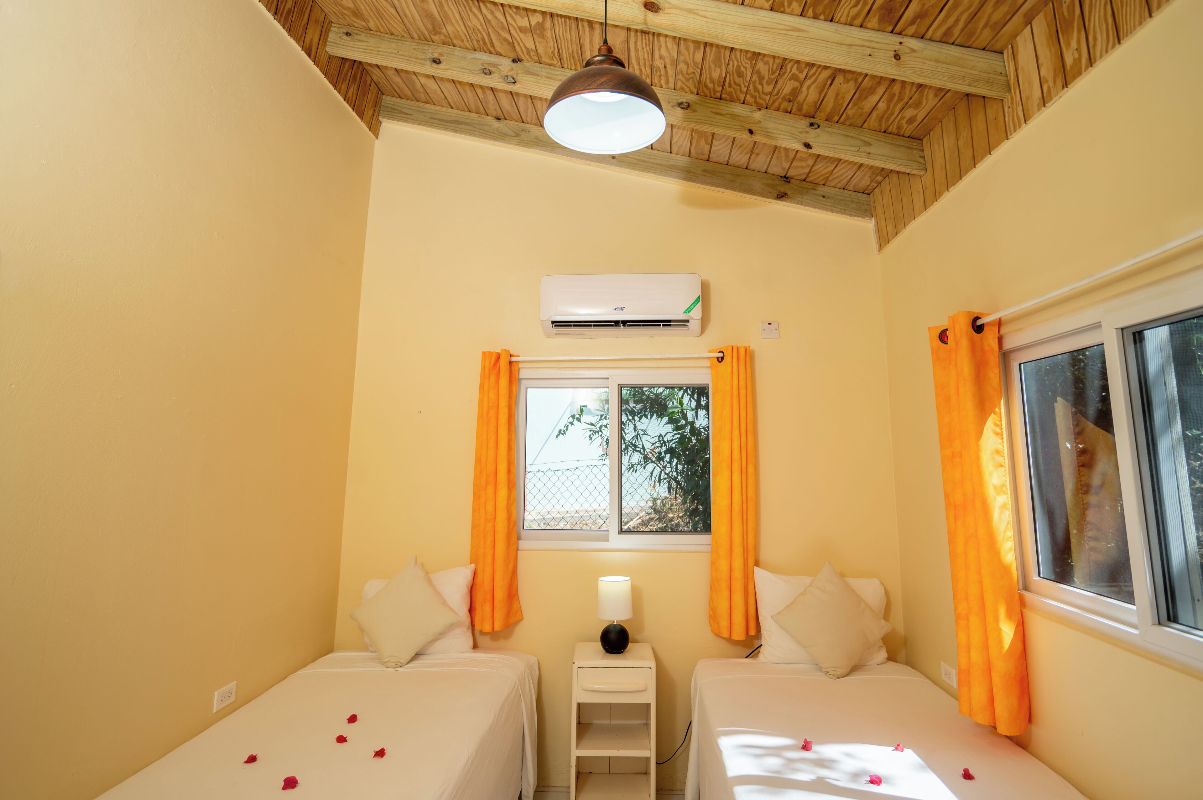 Una habitación con dos camas, aire acondicionado, ventana con cortinas y una mesita de noche con lámpara.
