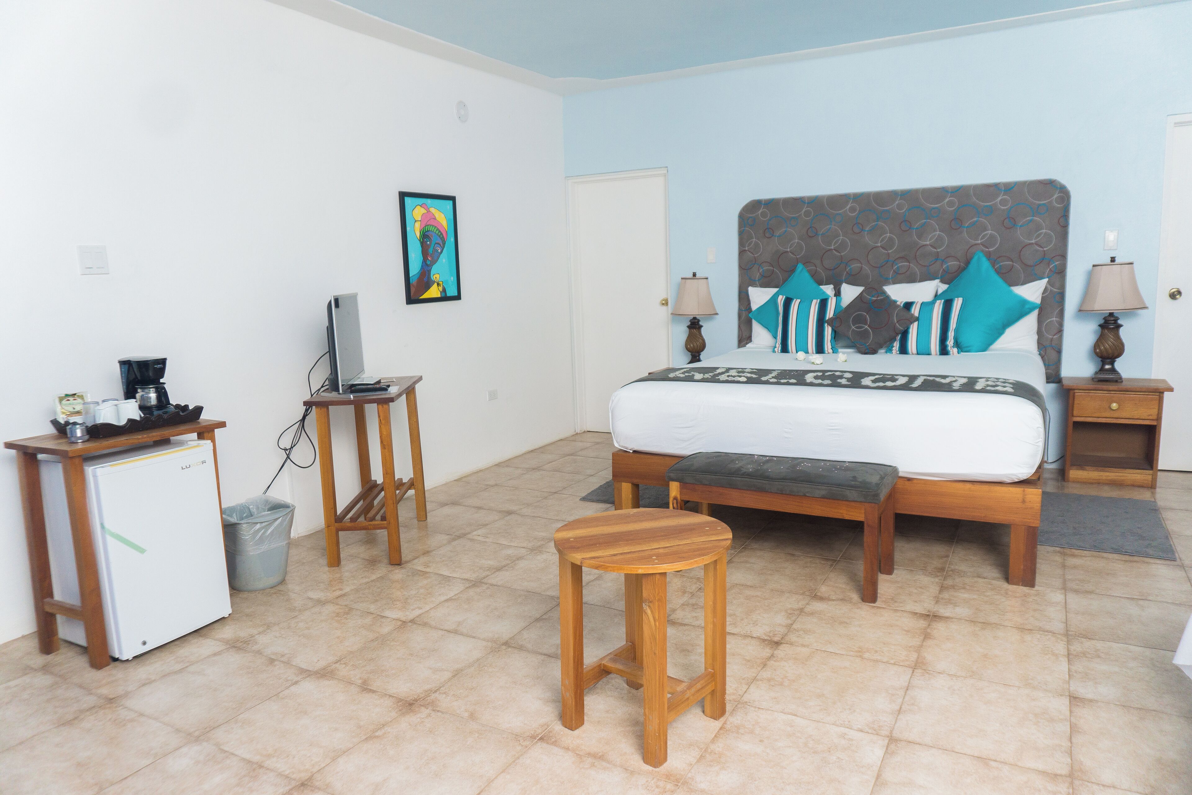 A bedroom with a bed, bedside tables, a desk, a refrigerator, and a trash bin.