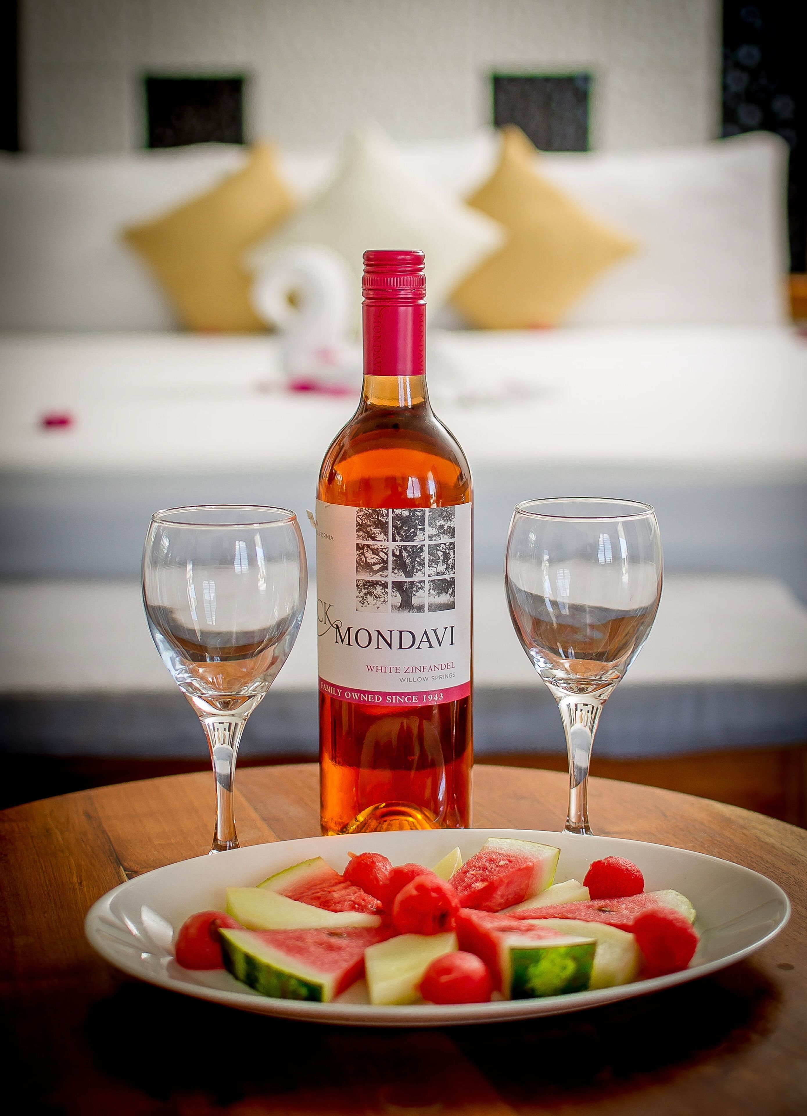 A bottle of Mondavi White Zinfandel on a table with two glasses and a plate of watermelon and raspberries.