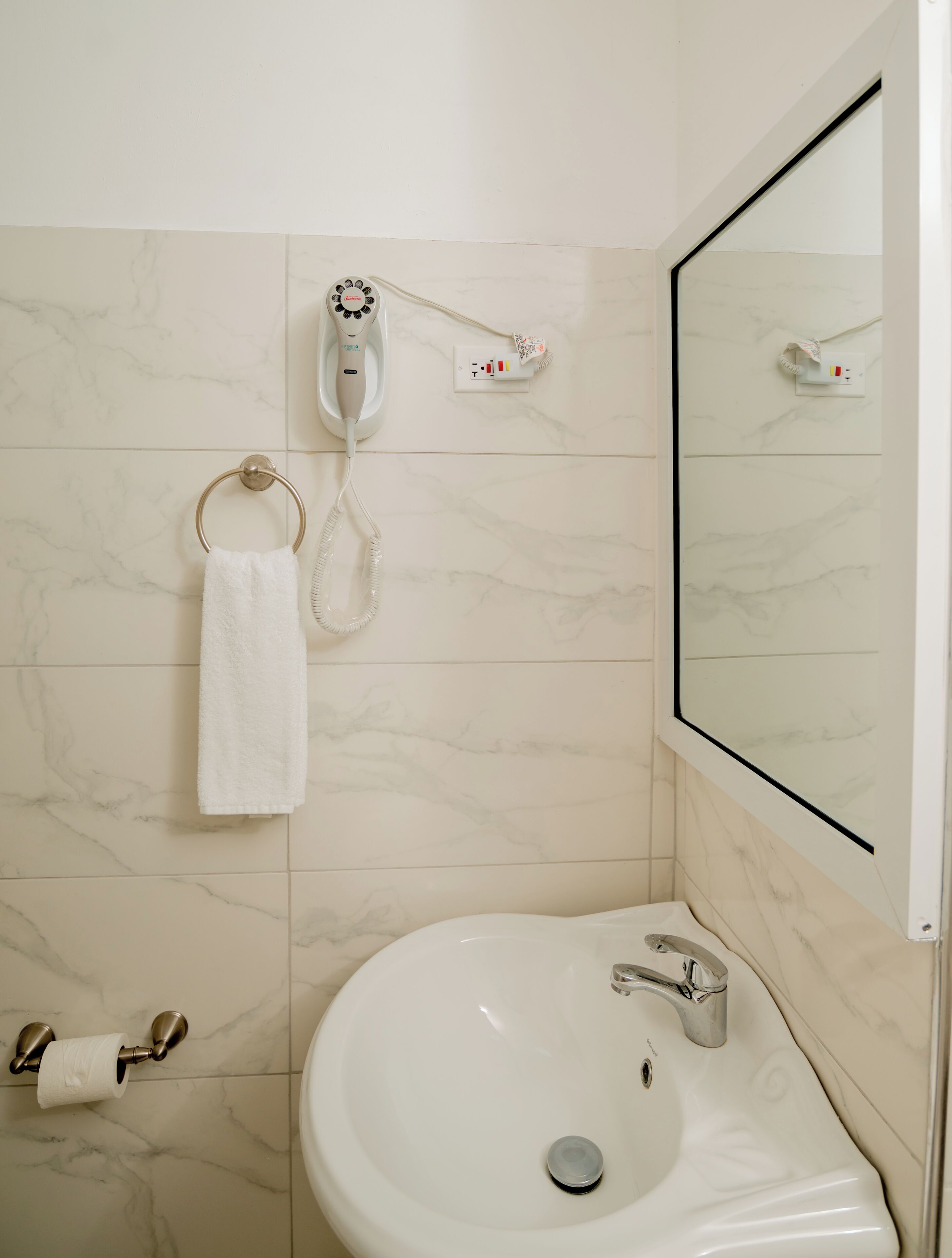 Een badkamer met een witte wastafel, een handdoekhouder, een toiletpapierrolhouder en een haardroger op een wandcontactdoos.