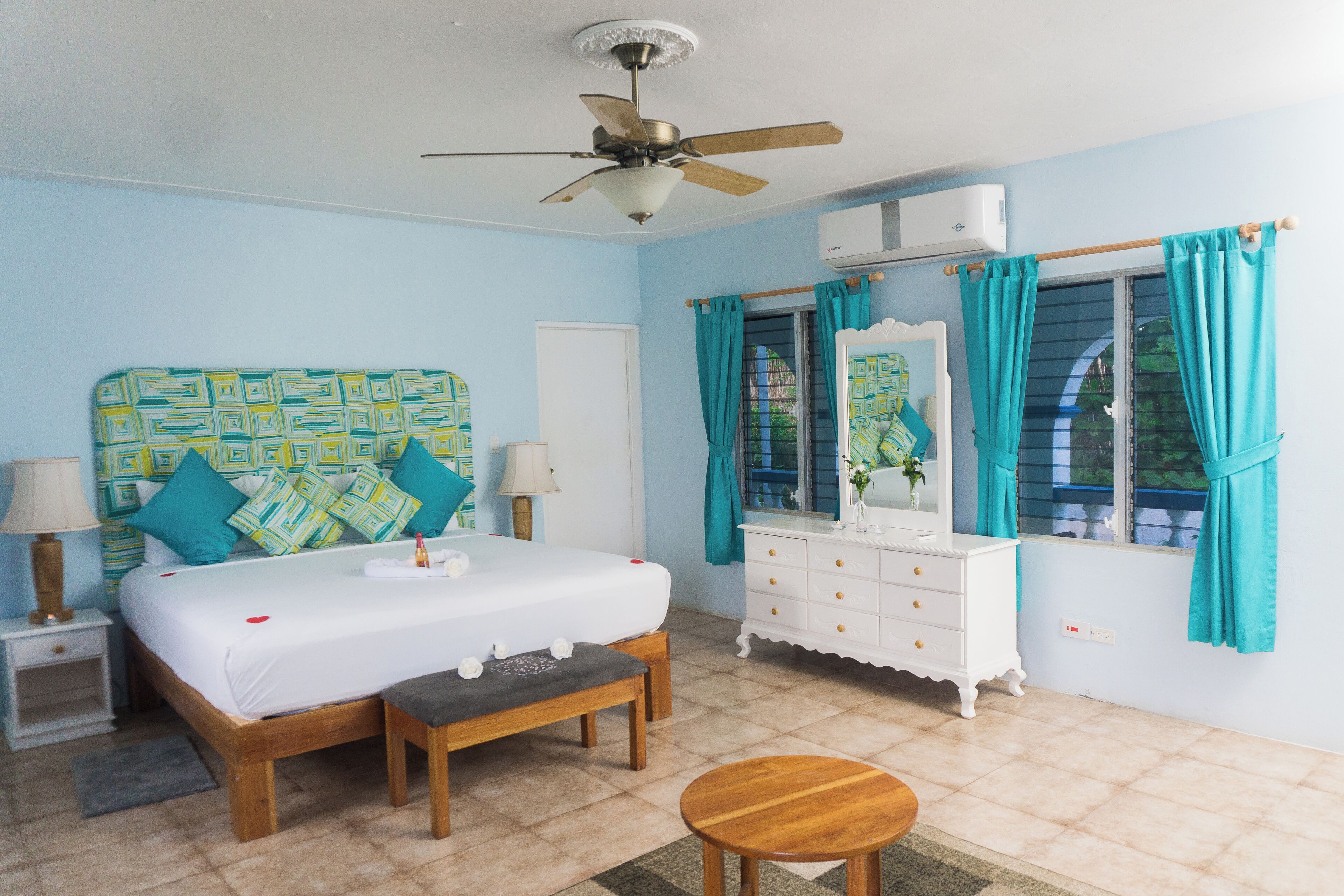 A bedroom with a bed, bedside tables, a dresser, a mirror, a ceiling fan, and a window with curtains.