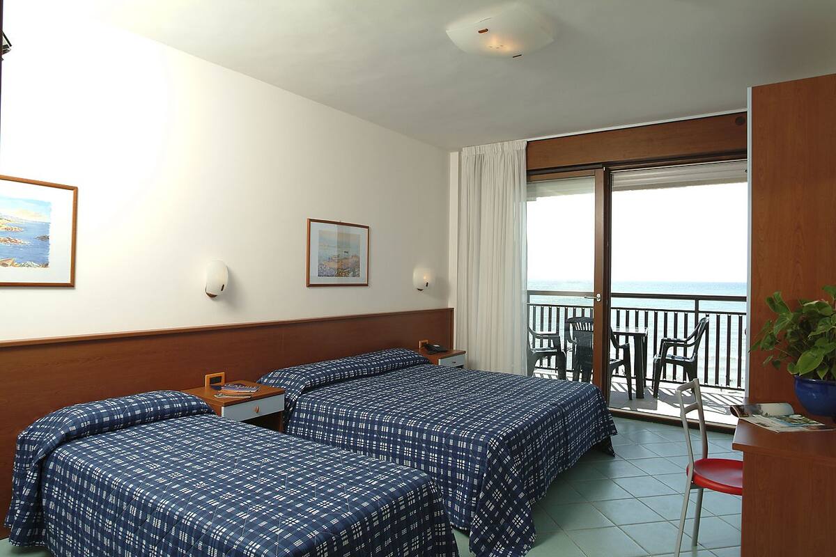 Habitación de hotel con dos camas, un escritorio, una silla, un balcón con vistas al mar y un cuadro en la pared.