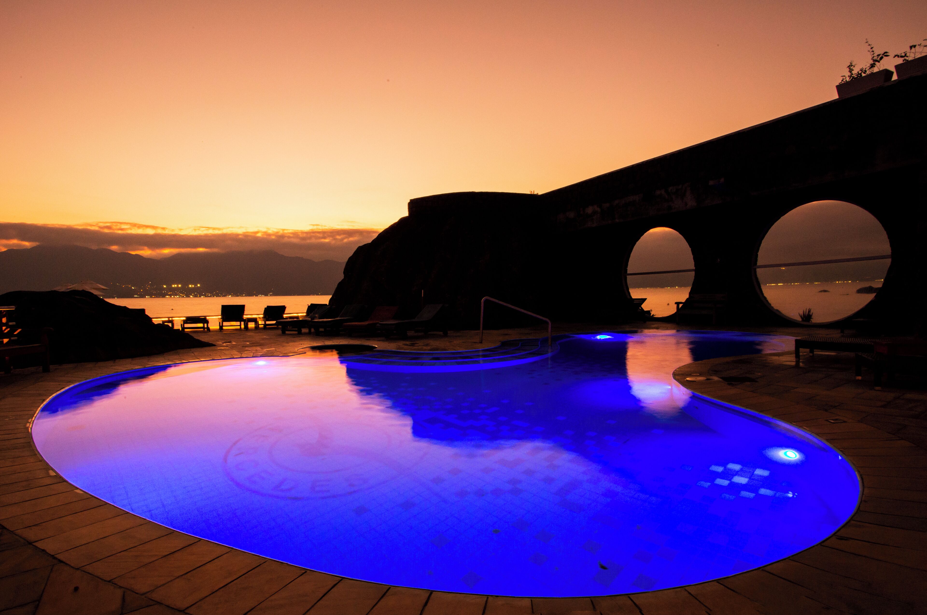 Una piscina iluminada con luces azules, un puente con arcos y una vista del agua y las montañas al fondo.