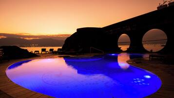 Una piscina iluminada con luces azules, un puente con arcos y una vista del agua y las montañas al fondo.