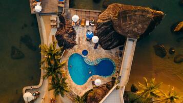 An aerial view of a resort with a long wooden pier, a swimming pool, and lounge areas with umbrellas.