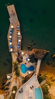 Vista aérea de un complejo turístico con un muelle de madera largo, una piscina y zonas de descanso con sombrillas.
