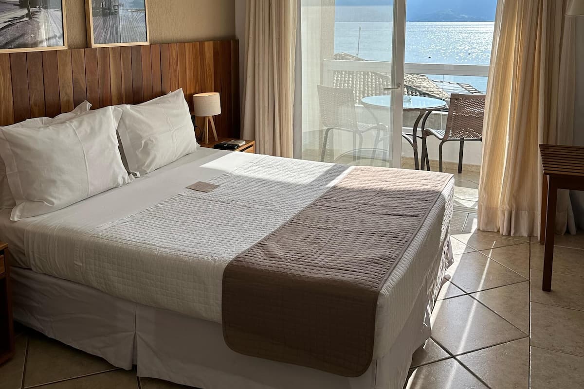 A hotel room with a bed, a ceiling fan, a large window with a view of the sea, and a balcony with outdoor furniture.