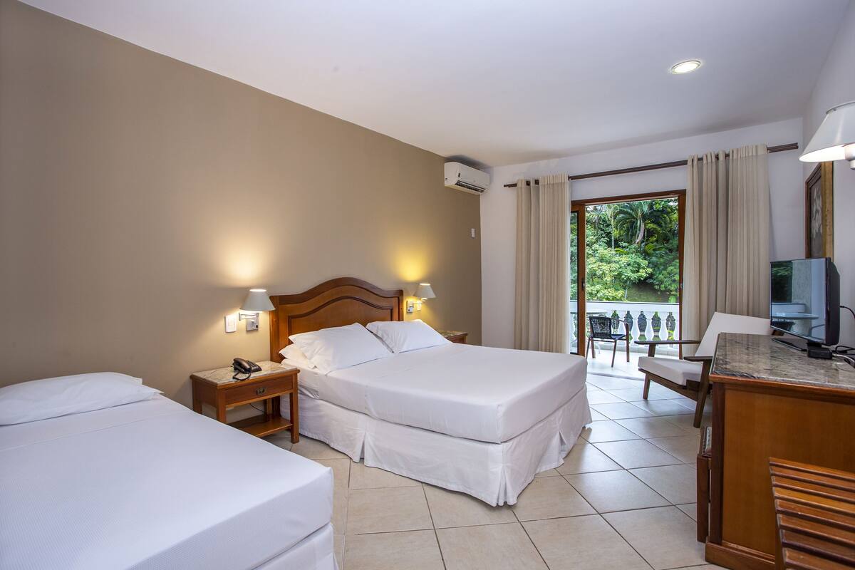 Habitación de hotel con dos camas, un escritorio con silla, televisión y balcón con vista a zonas verdes.