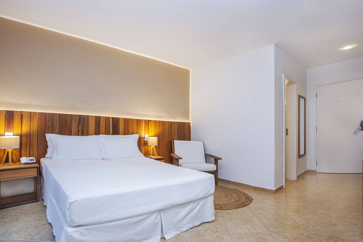Habitación de hotel con una cama grande, dos mesitas de noche con lámparas, una silla y un cabecero de madera.