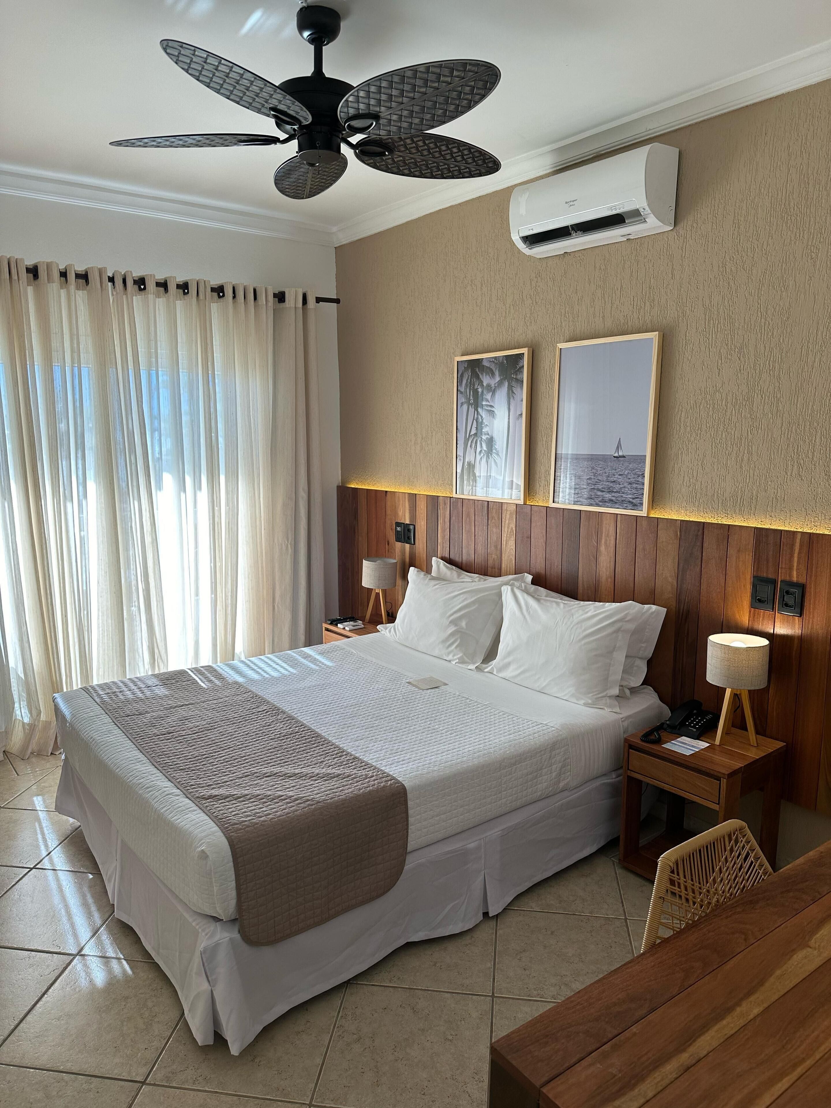 A hotel room with a large bed, a ceiling fan, an air conditioning unit, a wooden headboard, two framed pictures, a bedside lamp, a bedside phone, and a wooden table.