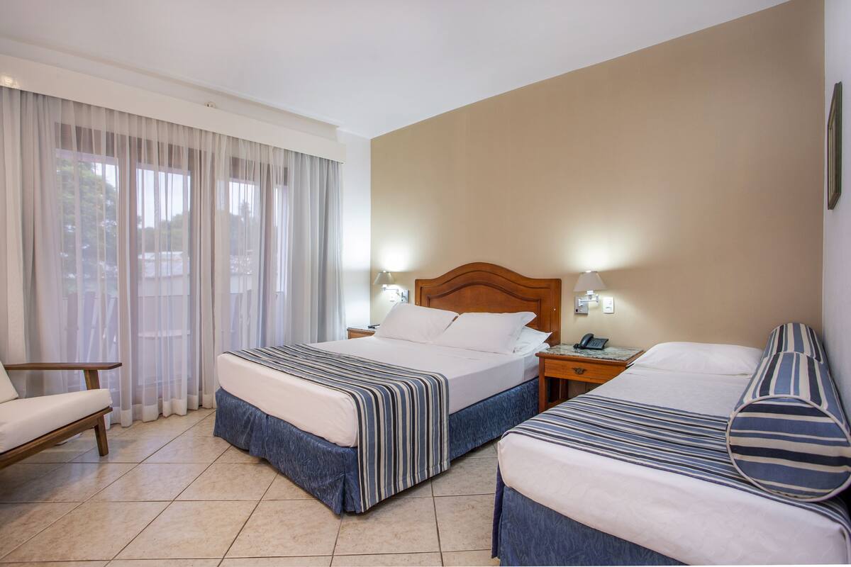 A hotel room with two beds, a chair, a nightstand with a lamp, and a window with sheer curtains.