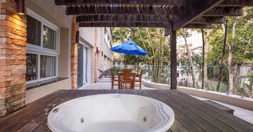 Un jacuzzi en una terraza de madera con una pérgola encima, una mesa y sillas, y un paraguas azul.