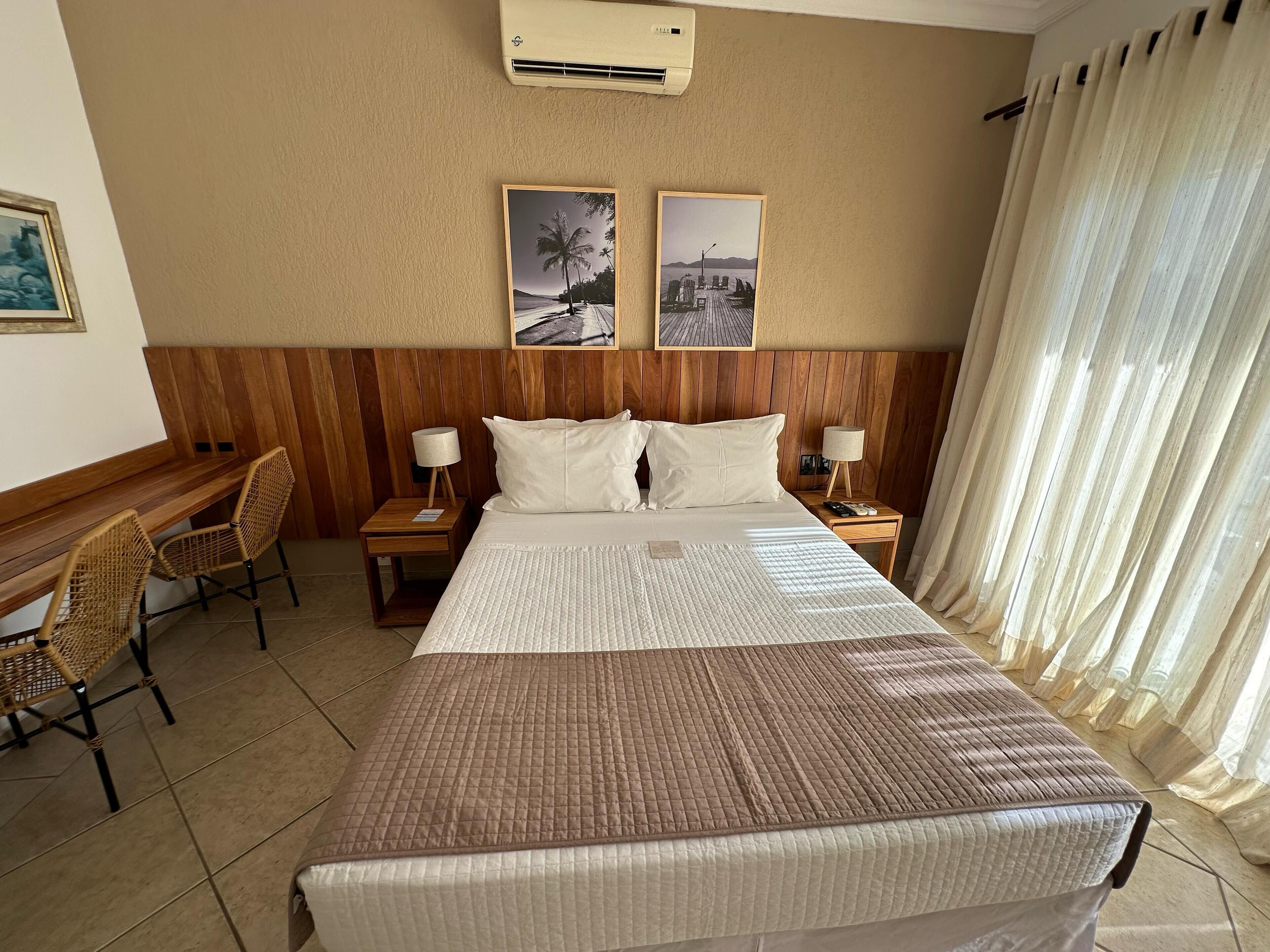Una habitación de hotel con una cama grande, dos mesitas de noche con lámparas, un escritorio de madera con sillas y dos cuadros en la pared.