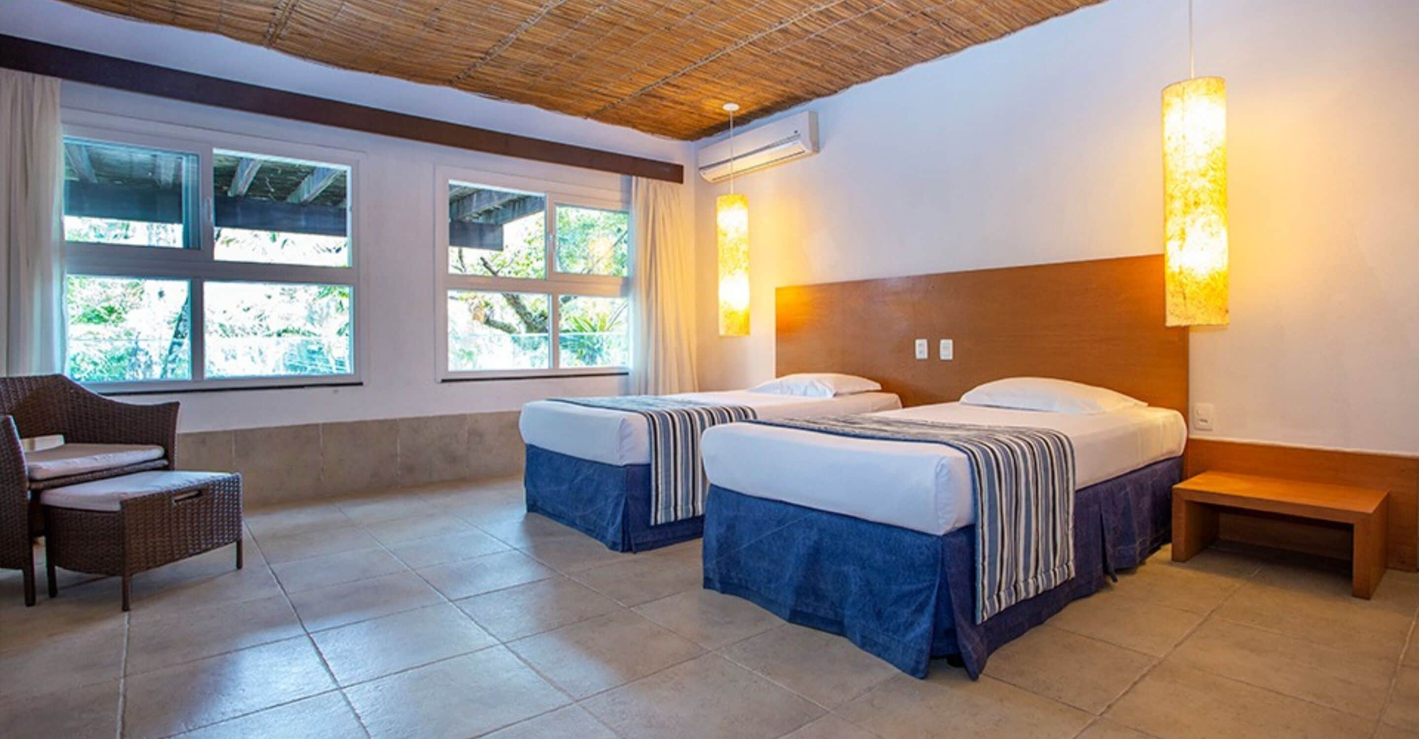 Ein Hotelzimmer mit zwei Betten, einem Sessel, einer Bank und großen Fenstern mit Vorhängen.