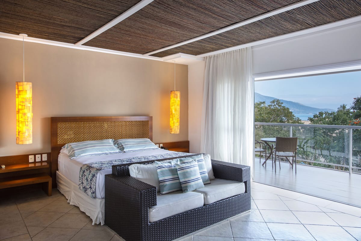 A bedroom with a large bed, a sofa, a balcony with a table and chairs, and a view of the outdoors.