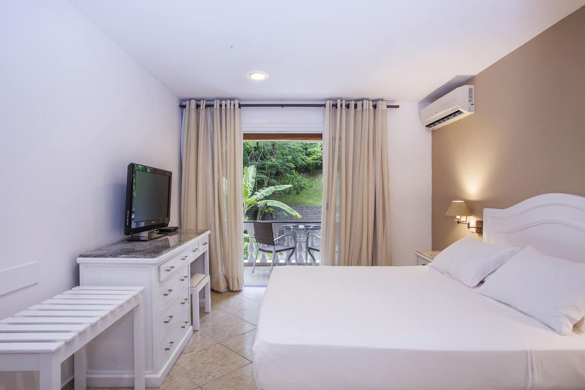 Habitación de hotel con cama, televisor en una cómoda, balcón con vistas a zonas verdes y unidad de aire acondicionado.