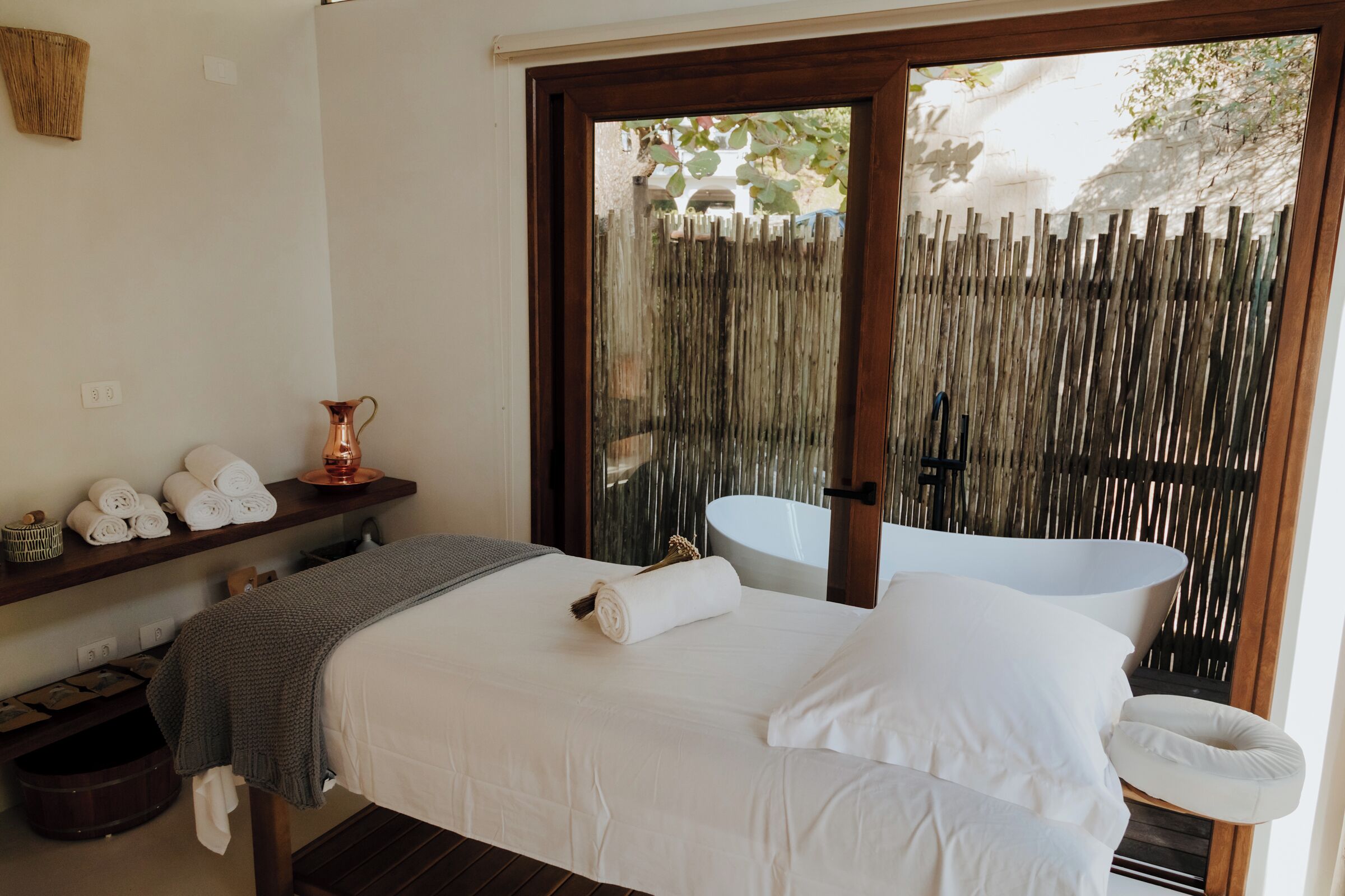 Une chambre à coucher avec un lit, une table de chevet avec des serviettes, un vase et une vue sur une clôture en bois et un évier à l’extérieur.