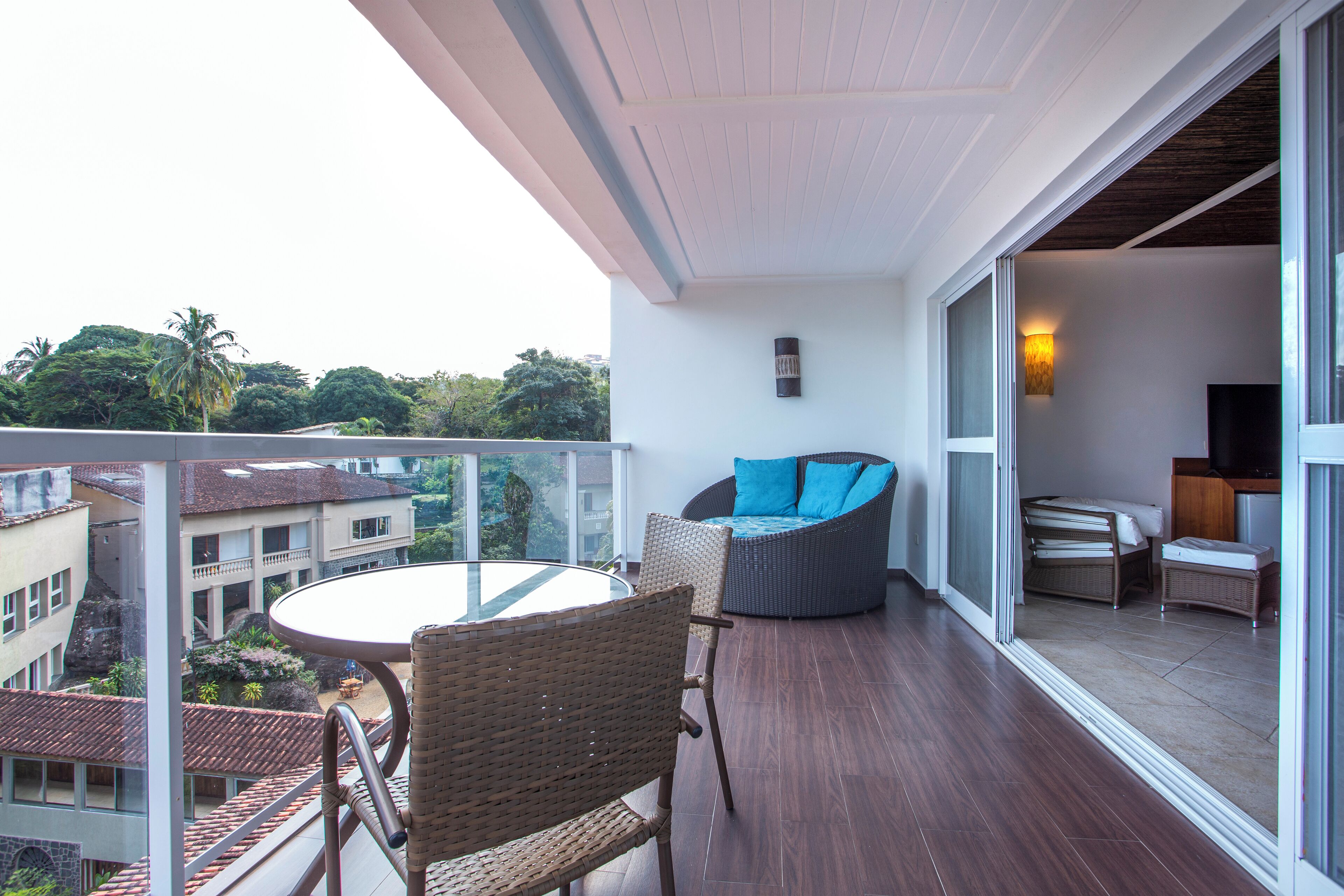 Un balcón con mobiliario de mimbre, una mesa de vidrio y vistas de los edificios y árboles circundantes.