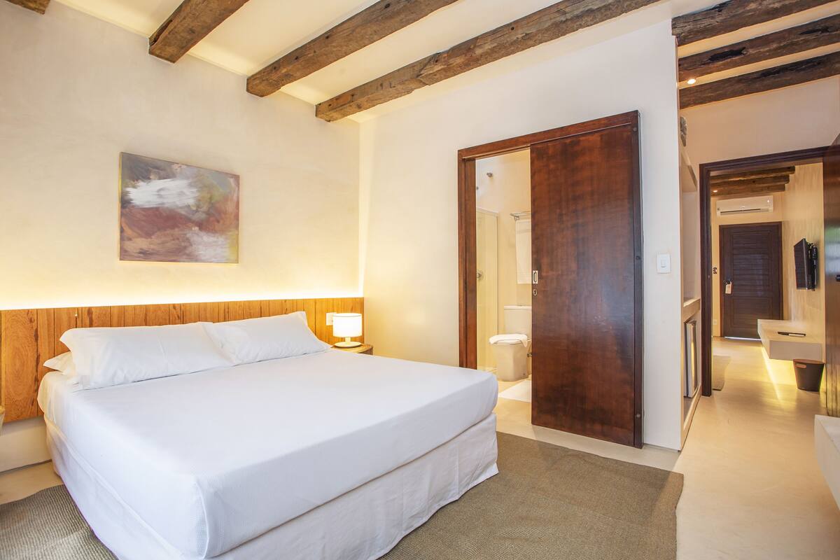 A hotel room with a large bed, wooden beams on the ceiling, a painting on the wall, and a bathroom visible through an open door.
