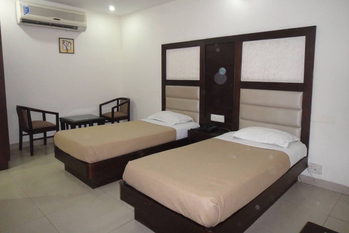 Habitación de hotel con dos camas individuales, una mesita, dos sillas, un equipo de aire acondicionado y un aplique de pared.
