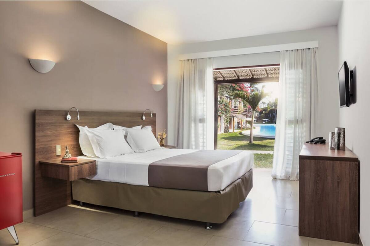 A hotel room with a bed, bedside tables, a TV, and a view of a swimming pool.