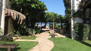 A pathway leading to a seating area with a thatched roof structure, surrounded by lush greenery and white walls.