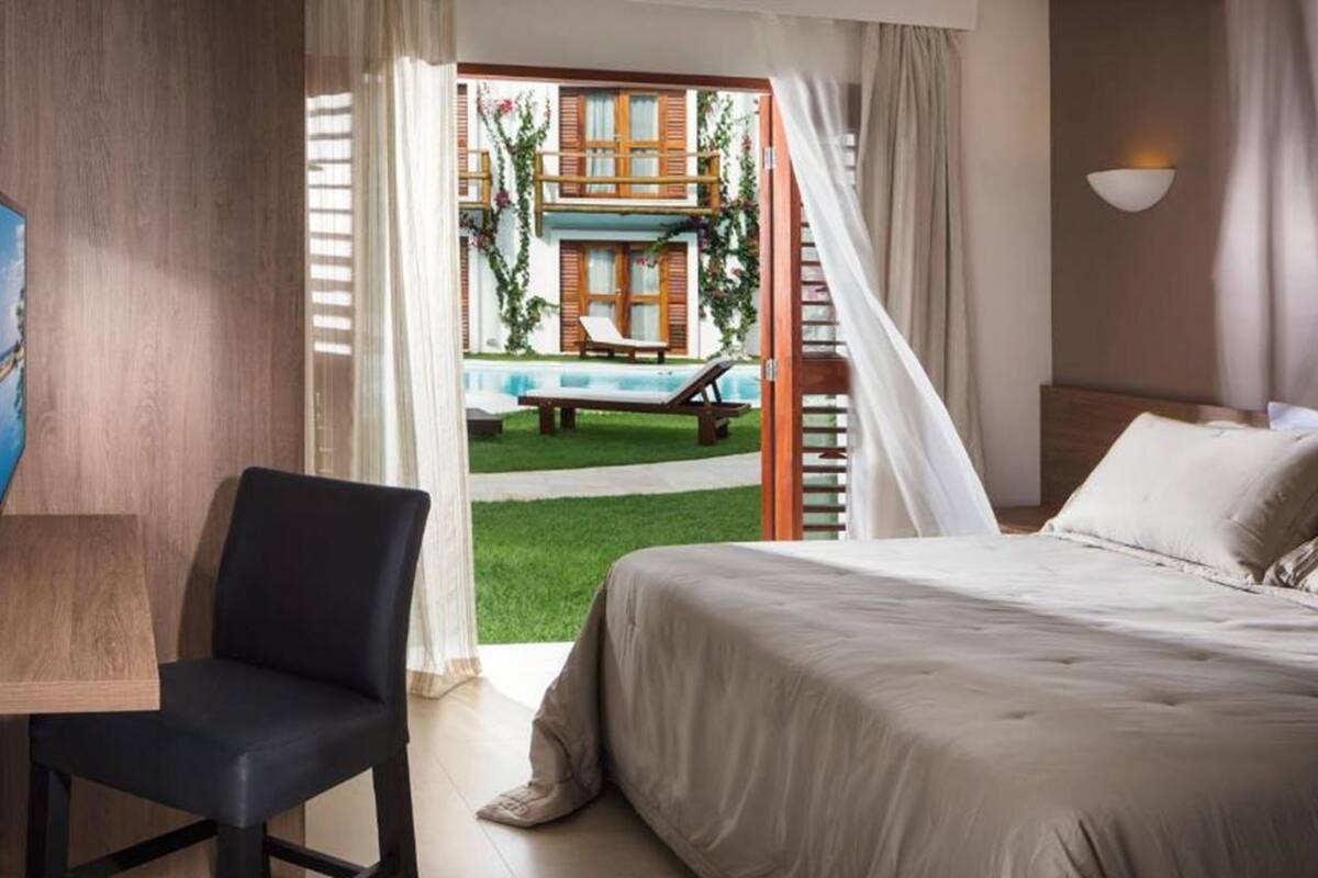 A hotel room with a bed, a chair, a painting, and a view of a pool and outdoor seating area.
