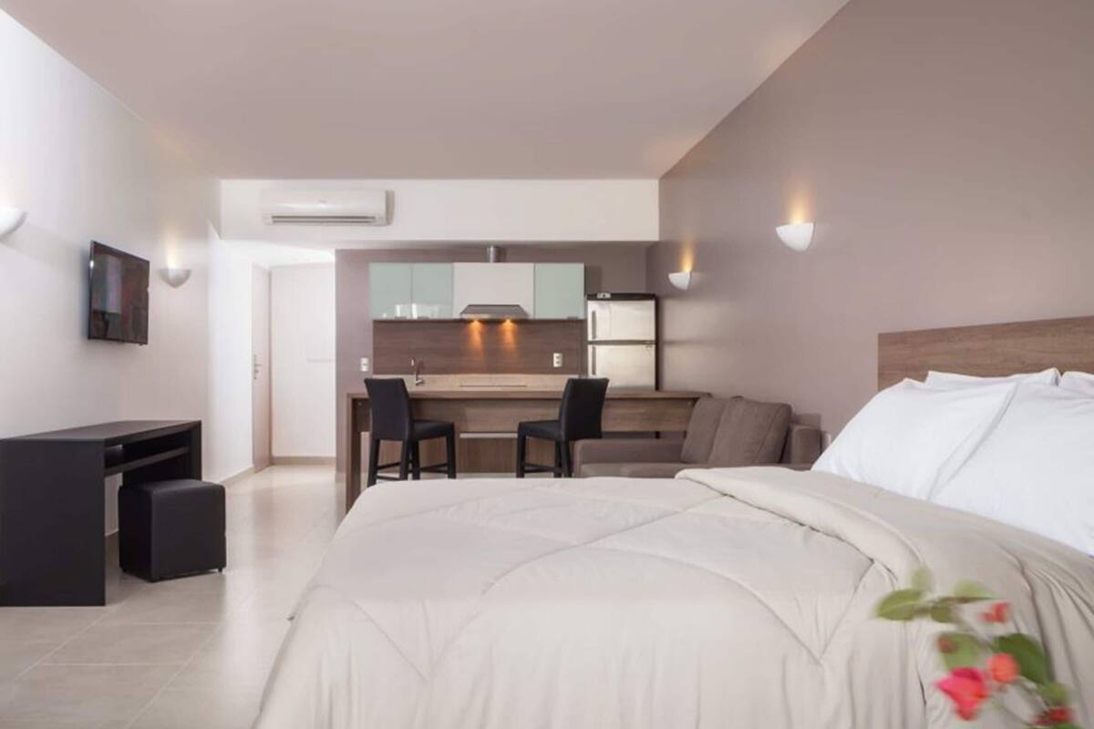 A modern hotel room with a large bed, a desk, a chair, a kitchenette, and a TV.