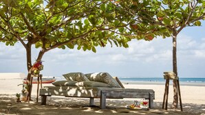 A beach setting with a wooden bench under a tree, a boat in the background, and a clear view of the ocean.