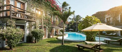 A pool area with lounge chairs and umbrellas, surrounded by lush greenery and a building with balconies.