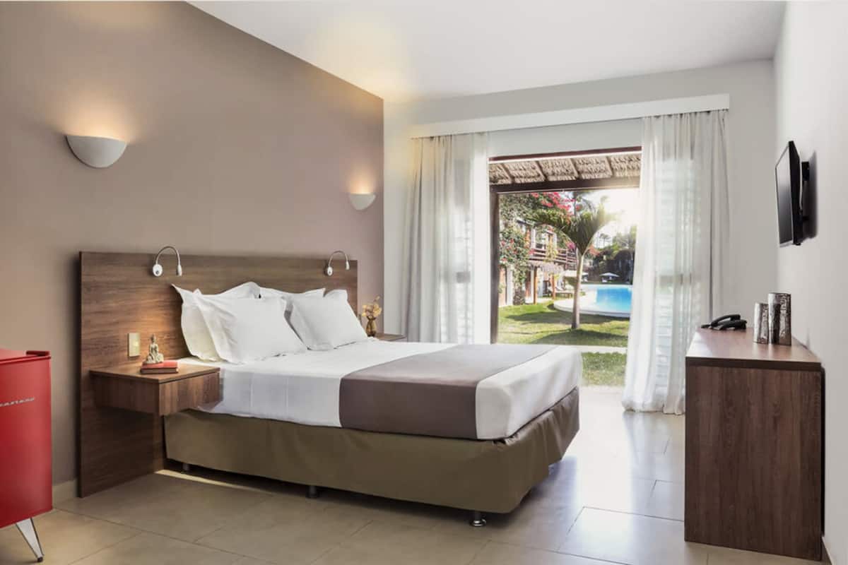 A modern hotel room with a large bed, bedside tables, a TV, and a view of a pool and garden.
