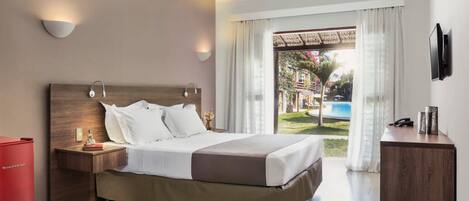 A modern hotel room with a large bed, bedside tables, a TV, and a view of a pool and garden.
