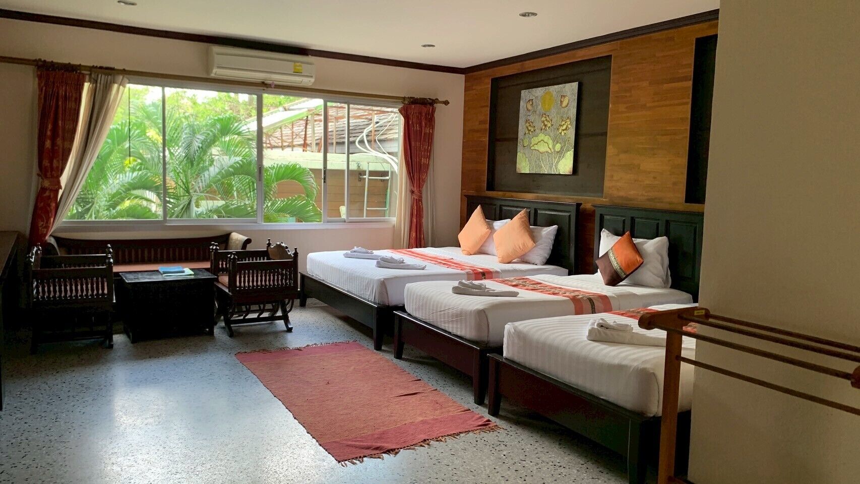 Habitación de hotel con dos camas, un pequeño rincón de descanso con un banco y una mesa, y un ventanal con cortinas.